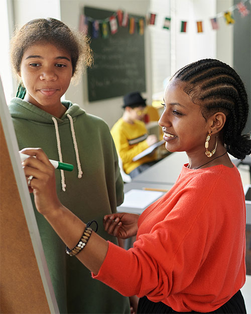 POC woman drawing on a whiteboard while a student watches and learns.
